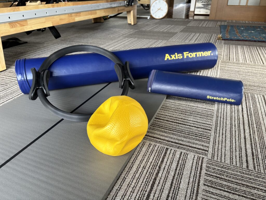 ピラティス用の小道具であるストレッチポール、ピラティスリング、黄色のソフトボールがマットの上に並べられている。
